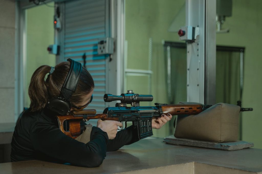 A woman in headphones aiming a sniper rifle at an indoor shooting range for target practice.